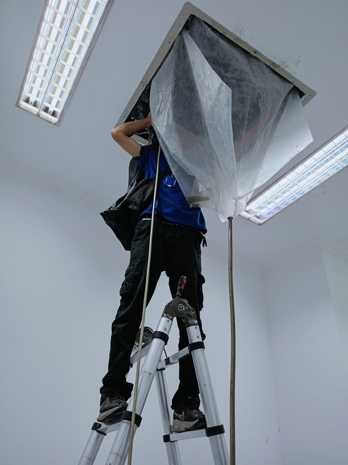 Teknisi melakukan cuci AC split wall di ruang kantor dengan plastik pelindung air